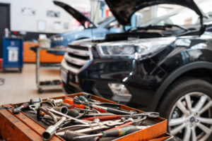 Various mechanical tools and instruments comprising screwdriver wrench plier placed in oily tray in front of car with opened bonnet and hanging headlight in service station of garage